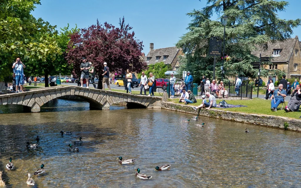 Stone footbridge over the River Windrush with ducks and visitors in Bourton-on-the-Water, Cotswolds