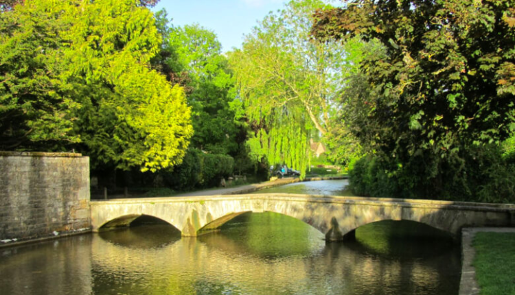 Stone bridge crossing the River Windrush in Burford surrounded by lush green trees on a sunny day