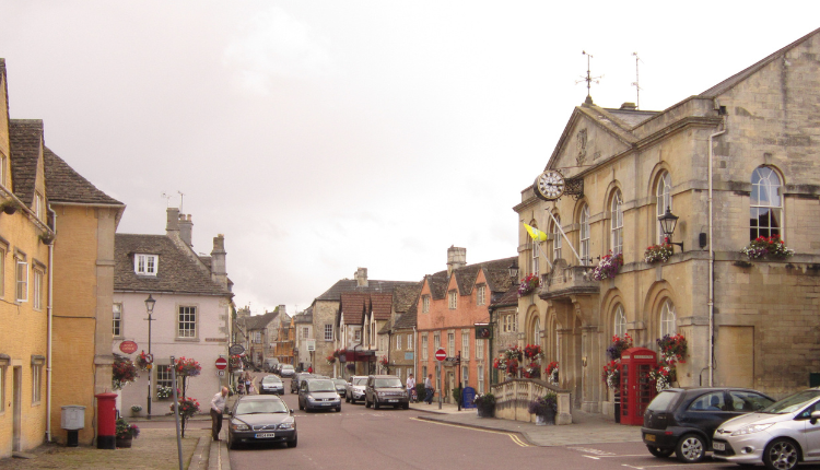 Corsham town centre with historic stone buildings, town hall, traditional shops, parked cars, and floral displays in Wiltshire