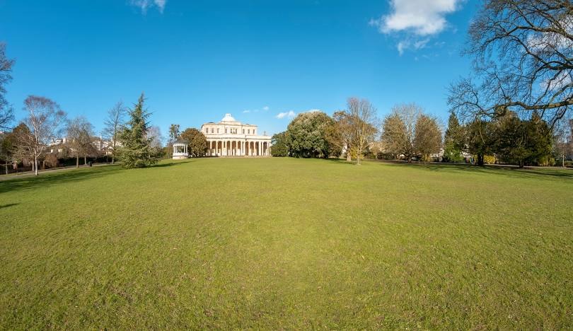 Pittville Pump Room in Cheltenham surrounded by open green parkland and trees.