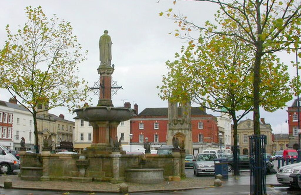 Stone monument and fountain in Devizes Market Square surrounded by historic buildings and trees