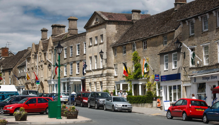 Historic High Street in Fairford with traditional Cotswold stone buildings, parked cars, and village shops.