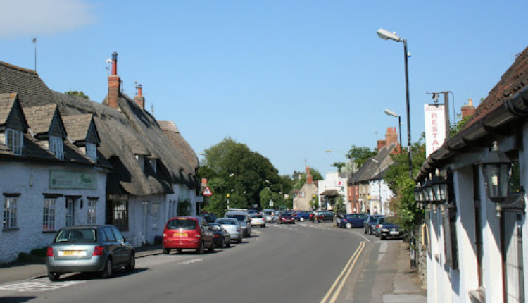 Street view of Shrivenham with traditional cottages, parked cars, and village shops along the road.