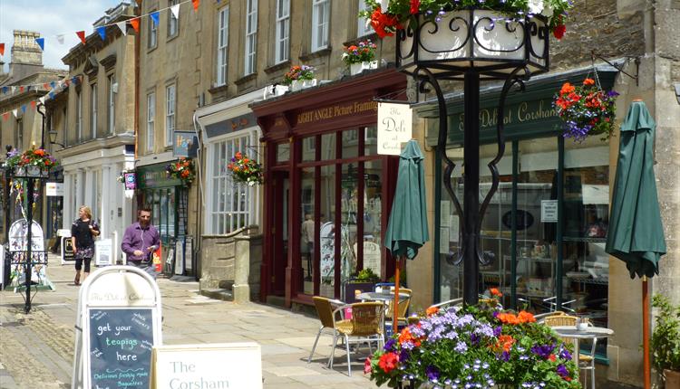 Corsham High Street with independent shops, outdoor café seating, and colourful hanging flower baskets