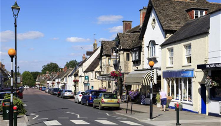 Cricklade High Street with historic Cotswold stone buildings, local shops, parked cars, and hanging flower baskets.